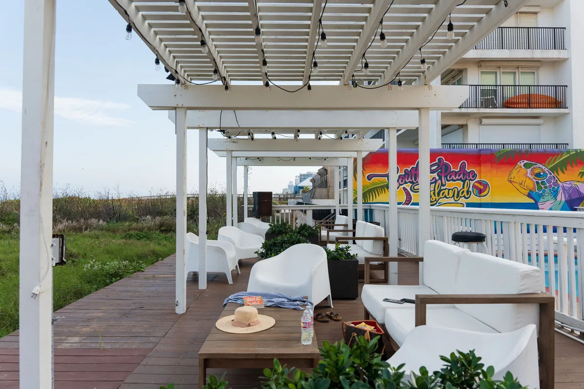 A boardwalk lined with white lounge seating under pergolas, with the colorful South Padre Island mural behind