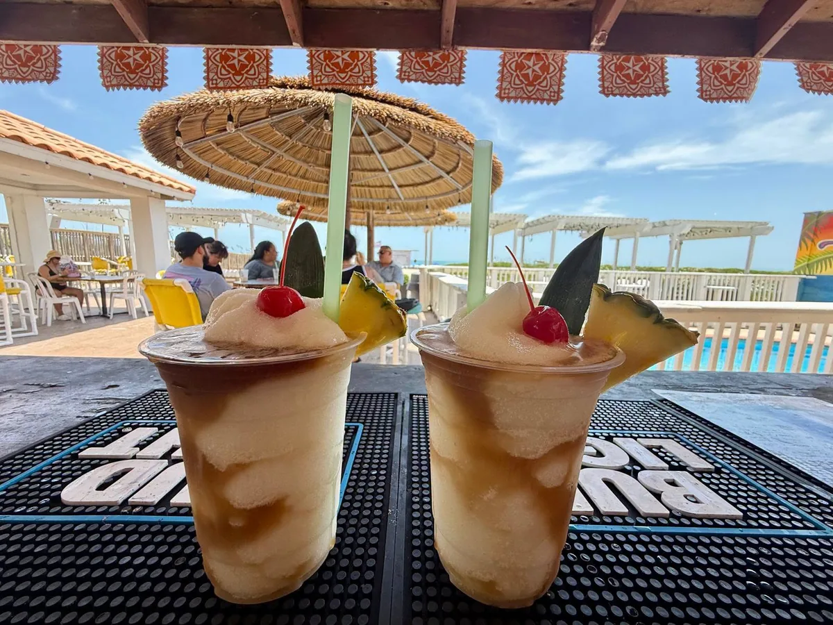 Two frozen drinks topped with cherries and pineapple on the bar, with the thatched umbrellas and the deck behind