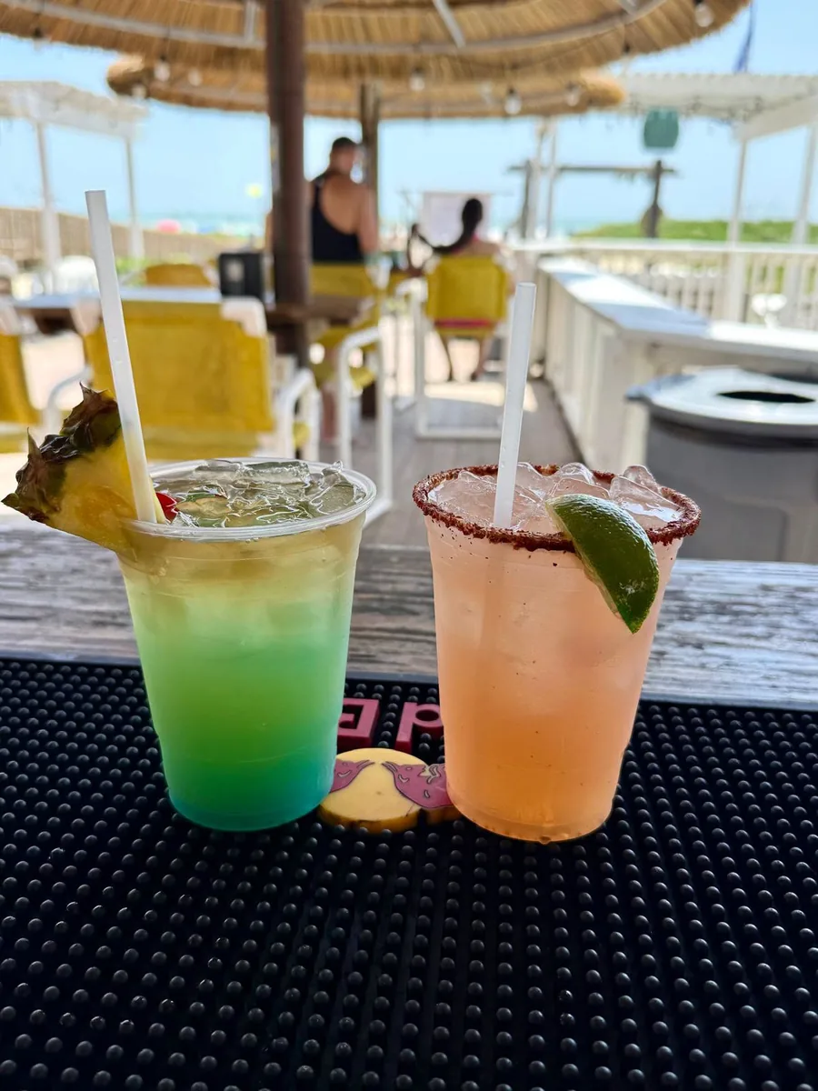 A green and blue layered drink with pineapple and an orange drink with a chili-salt rim and lime, on the bar