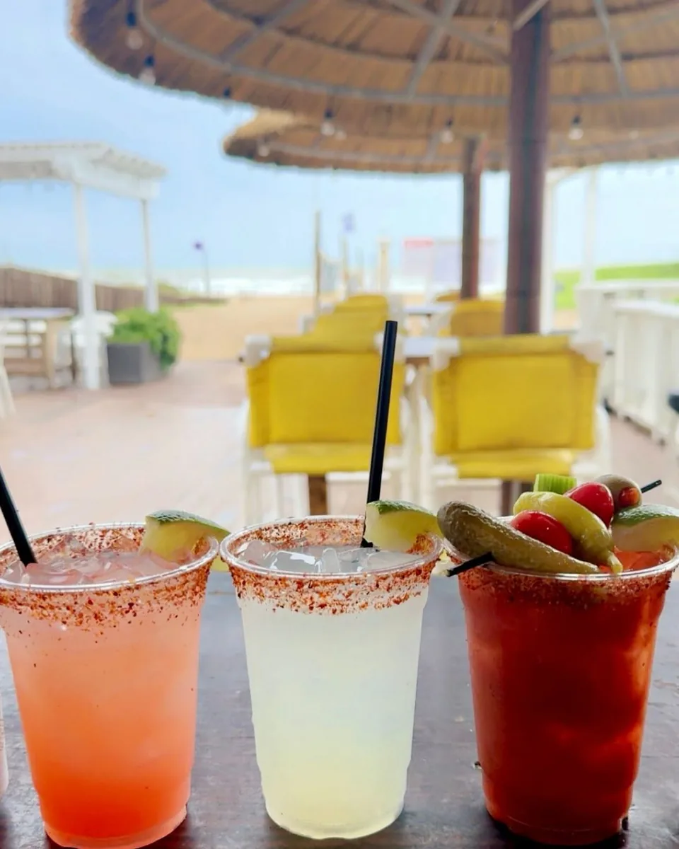 Three drinks on the bar: a pink drink and a margarita with chili-salt rims, and a red michelada with a skewer of fruit and pickles