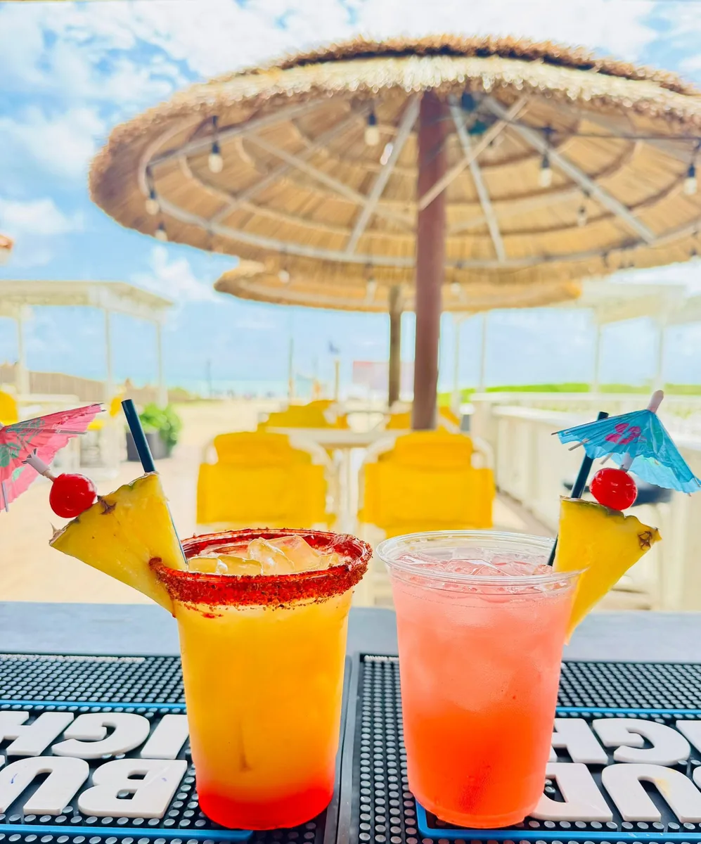 Two tropical cocktails with paper umbrellas, pineapple and cherries, one with a chili-salt rim, on the bar under a thatched umbrella by the beach