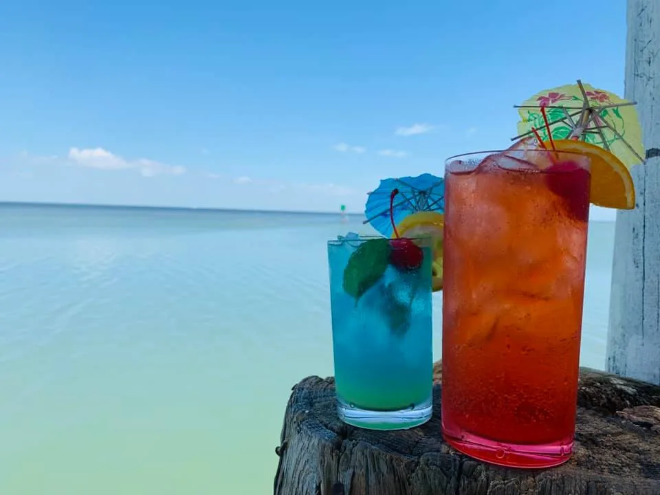 A blue cocktail and a red cocktail with fruit and paper umbrellas on a post above the bay