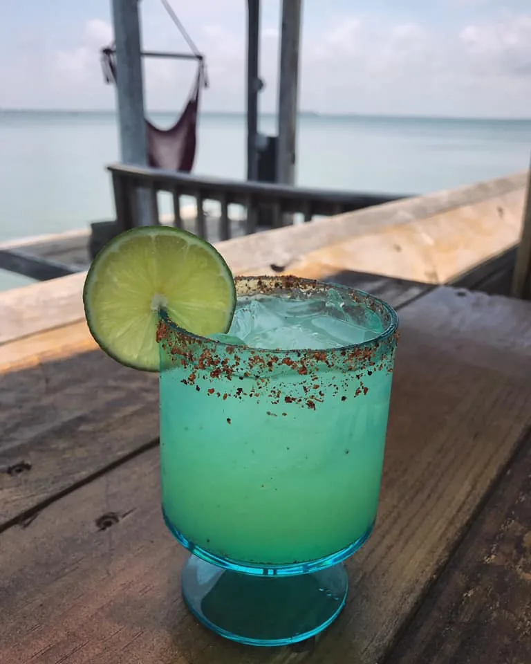 A turquoise cocktail with a chili-salt rim and a lime wheel on a wooden table by the water