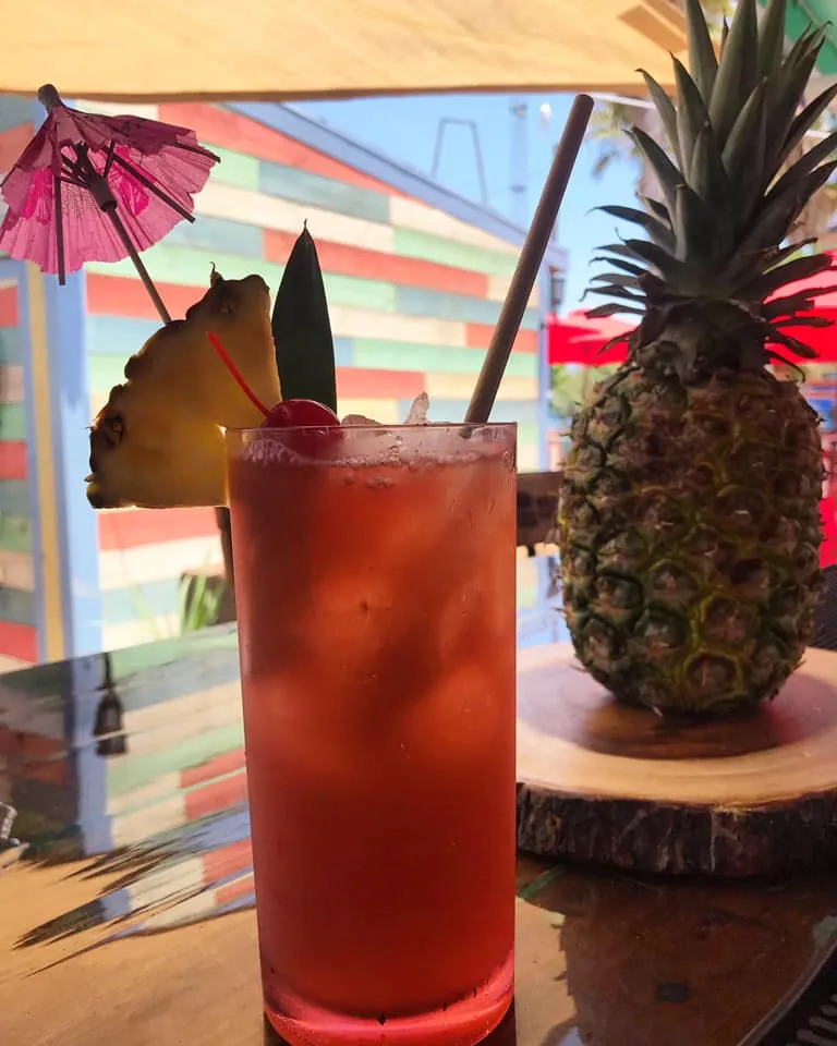 A red cocktail with a pineapple wedge, a cherry and a paper umbrella next to a whole pineapple at the bar