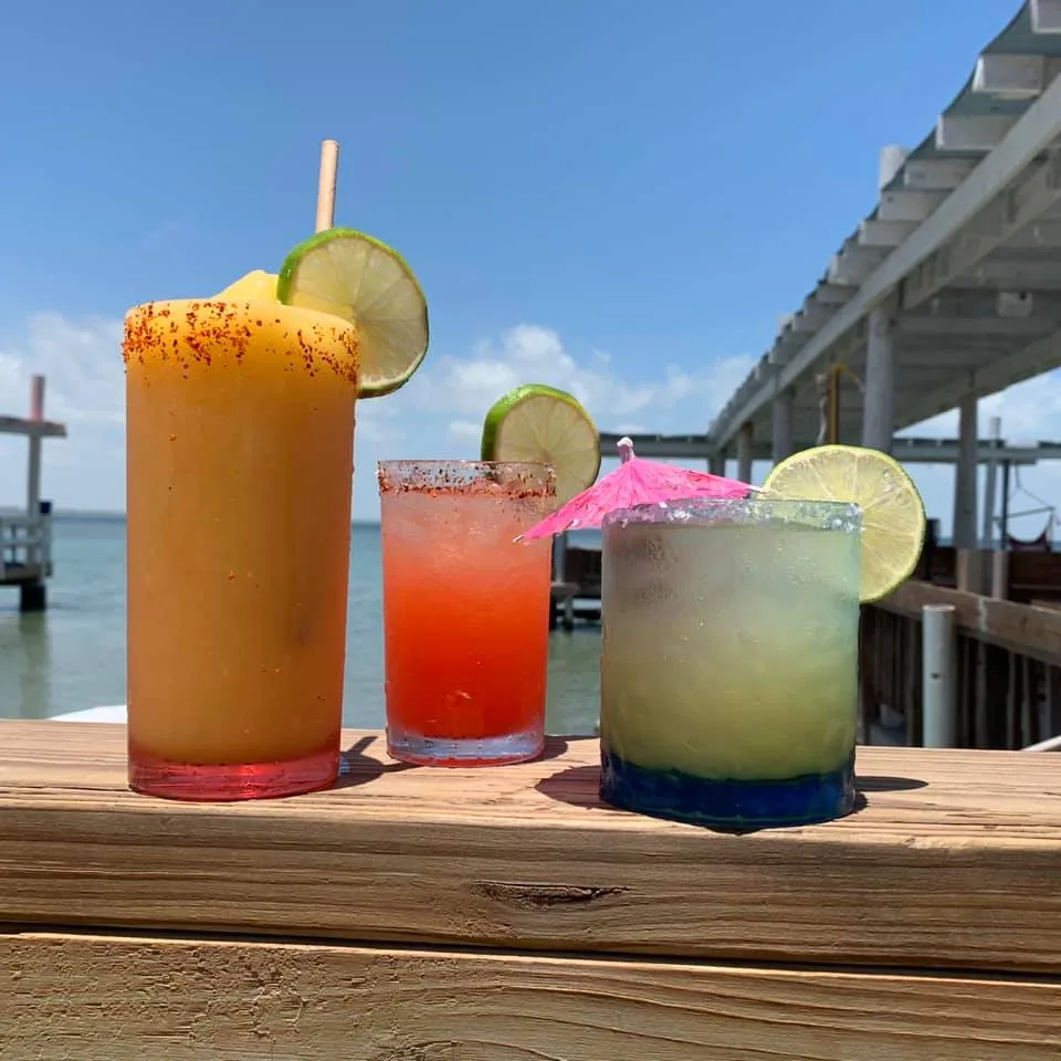 Three cocktails with chili-salt rims and lime wheels lined up on the deck rail over the water