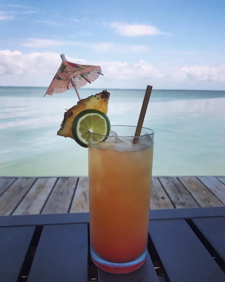 An orange cocktail garnished with pineapple, a lime wheel and a paper umbrella, on the deck rail over the bay