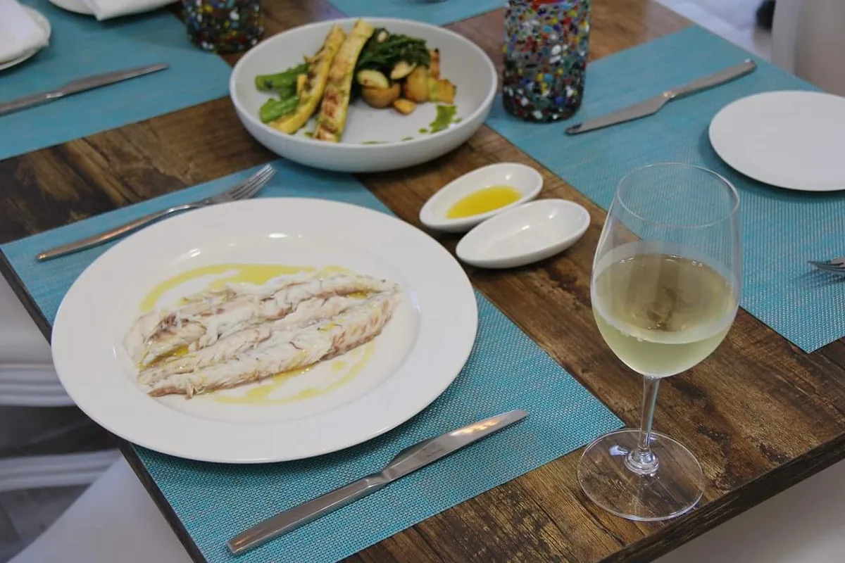 Fish fillets dressed with olive oil on a white plate, with grilled vegetables, lemon oil and a glass of white wine