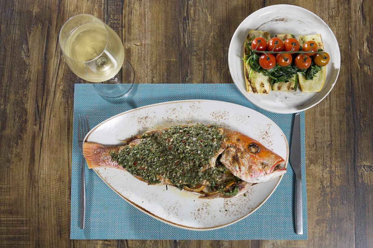 A whole red snapper under a green herb topping on an oval platter, with a side of tomatoes and greens and a glass of white wine