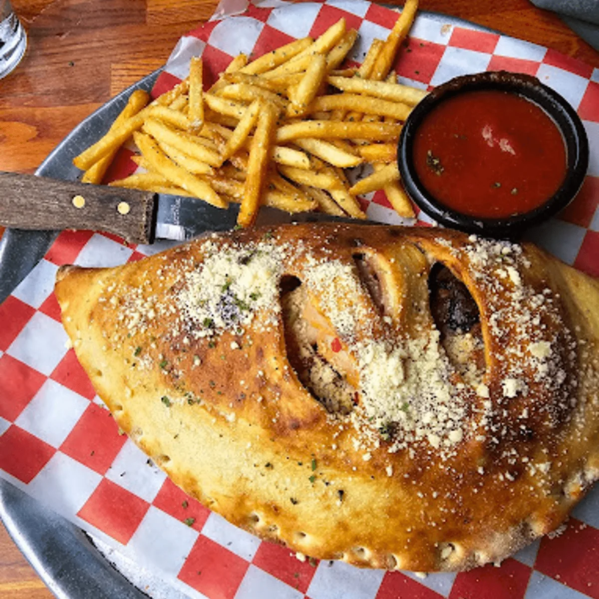 A golden brick-oven calzone dusted with Parmesan, with fries and a cup of marinara on checkered paper