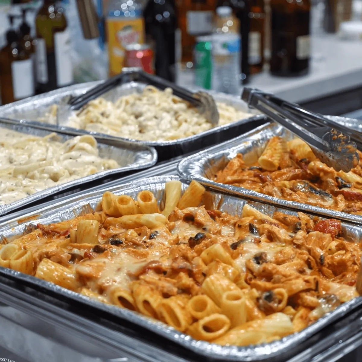Foil catering pans of Alfredo pasta and rigatoni in a red cream sauce, with serving tongs