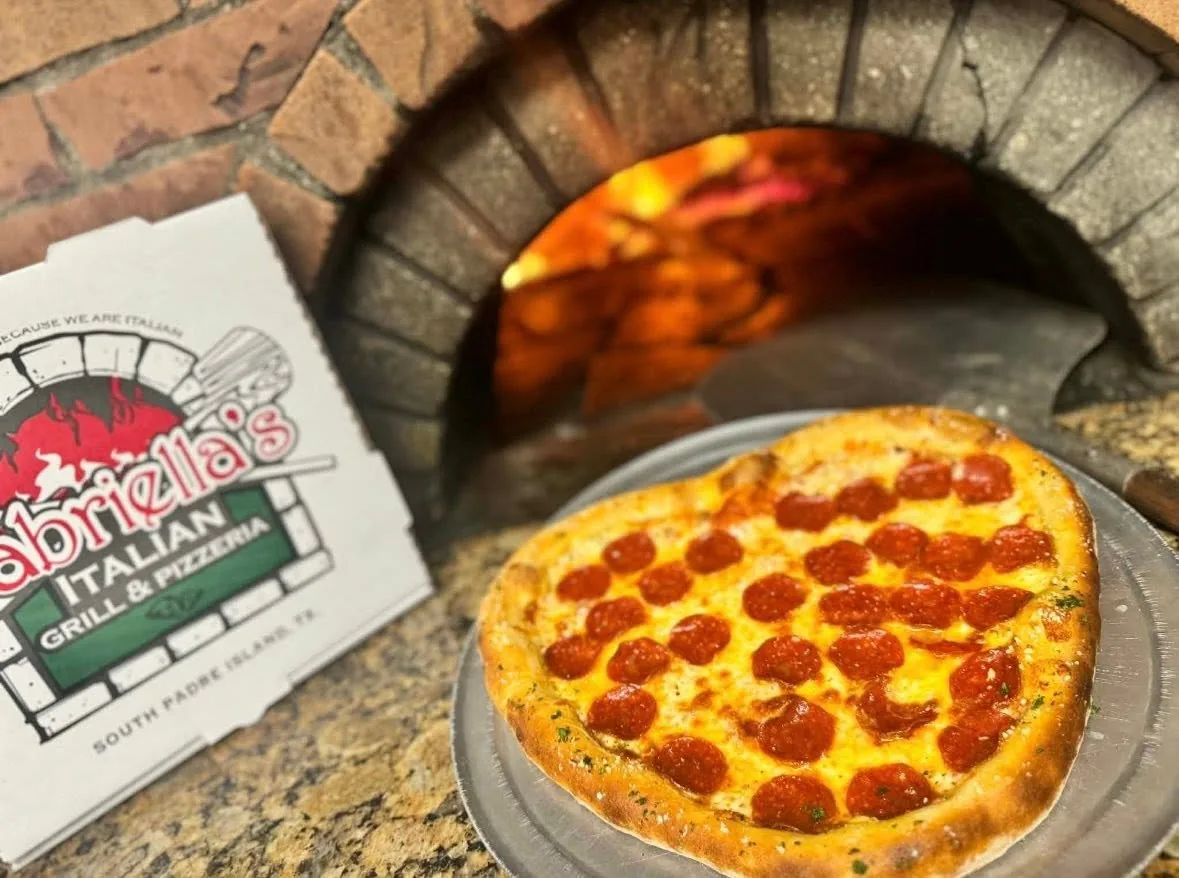 A heart-shaped pepperoni pizza on a metal tray in front of the brick pizza oven, next to a Gabriella's pizza box