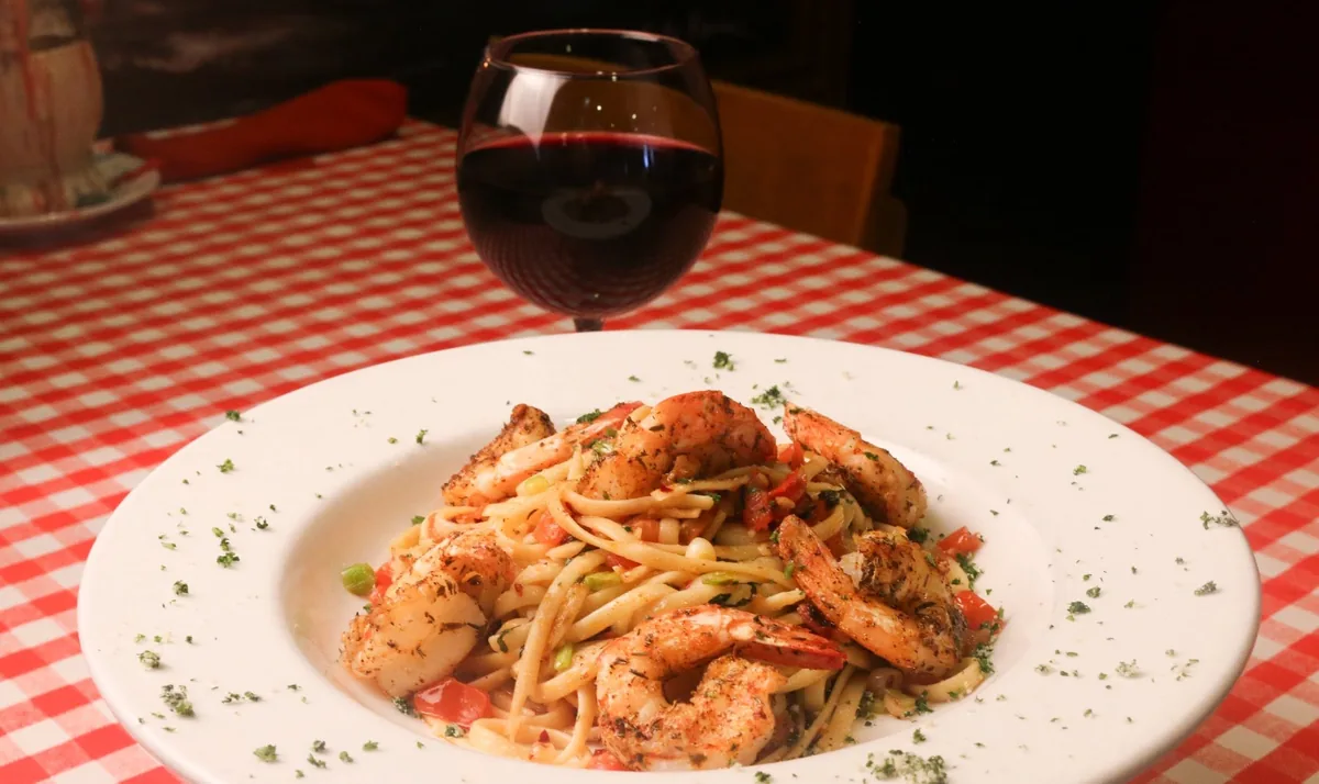 Seasoned shrimp over linguine with diced tomatoes and green onions, next to a glass of red wine on a red-checked tablecloth