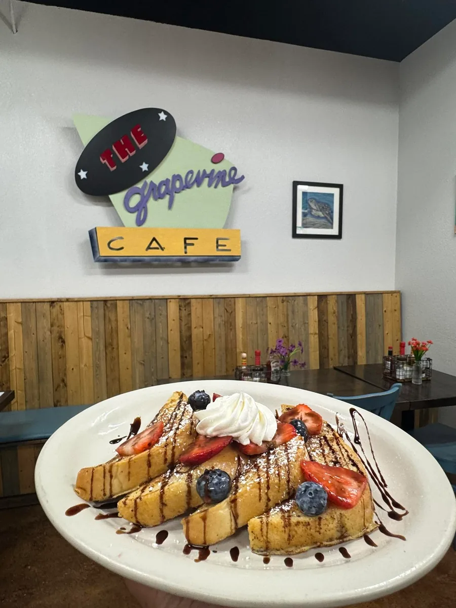 A plate of French toast with berries and whipped cream held up in front of the Grapevine Cafe sign in the dining room