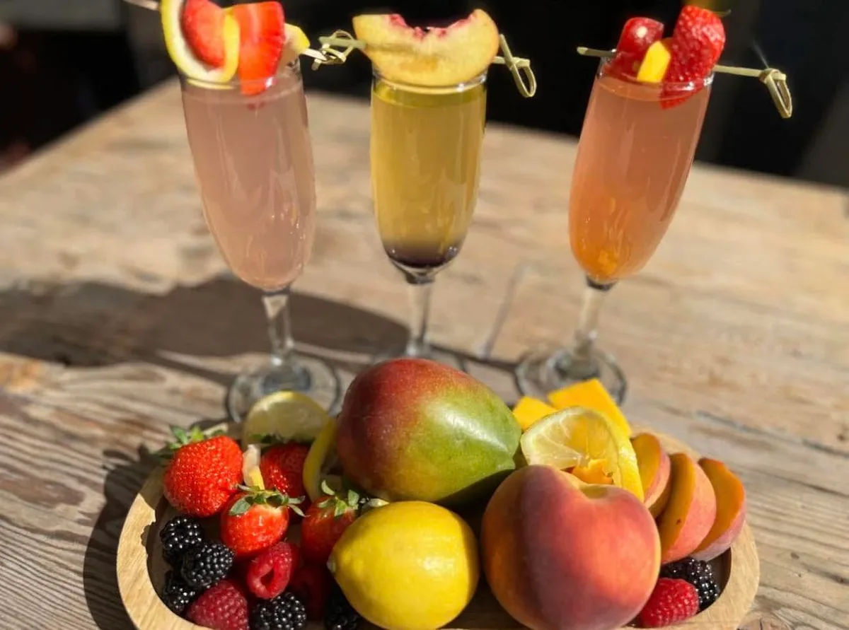 Three fruit mimosas in champagne flutes with a board of strawberries, mango, peaches and lemons