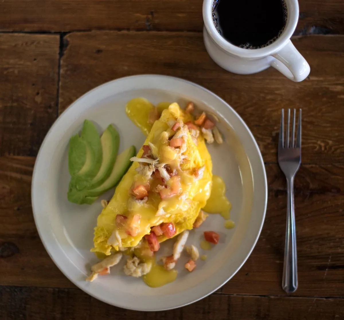 An omelette topped with hollandaise and diced tomato, with avocado slices and a cup of coffee