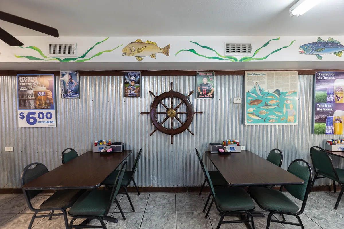 The dining room with tables along a corrugated-metal wall hung with a ship's wheel and a painted fish border