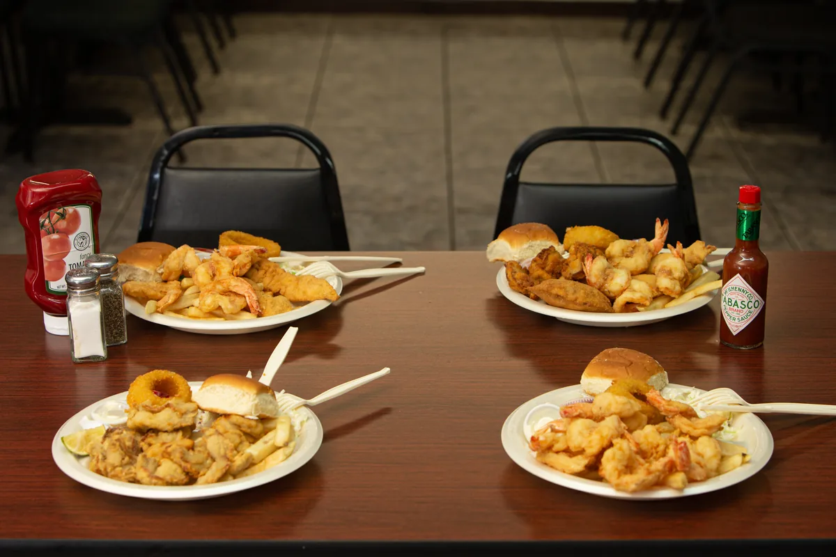 Four plates of fried shrimp, fish and oysters with rolls on a table, with ketchup, hot sauce and salt and pepper