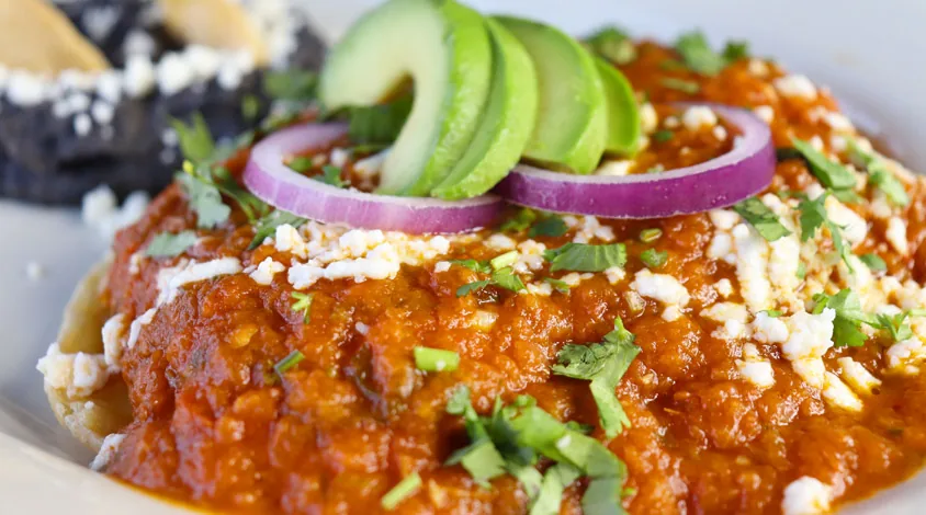 Close-up of red chilaquiles topped with queso fresco, red onion rings, avocado slices and cilantro