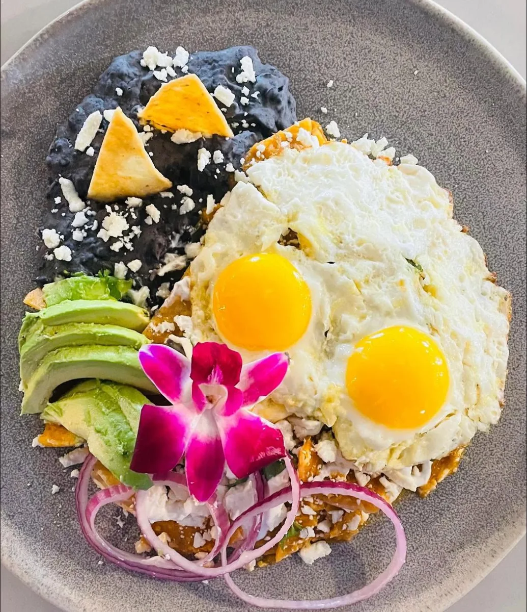 Red chilaquiles with two fried eggs, black beans with tortilla chips, avocado, red onion and an orchid on a grey plate