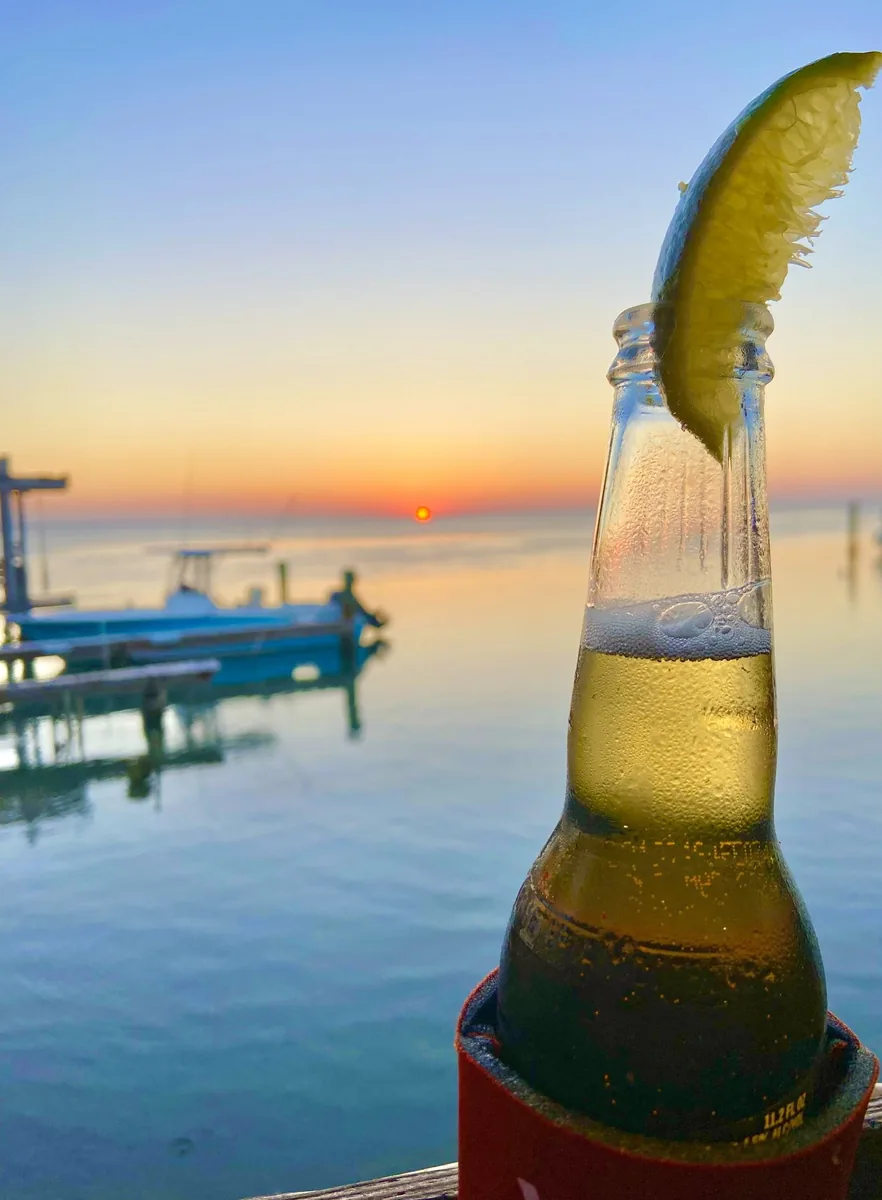 A bottle of beer with a lime wedge on the deck rail, with boats and the sun setting over the bay behind it