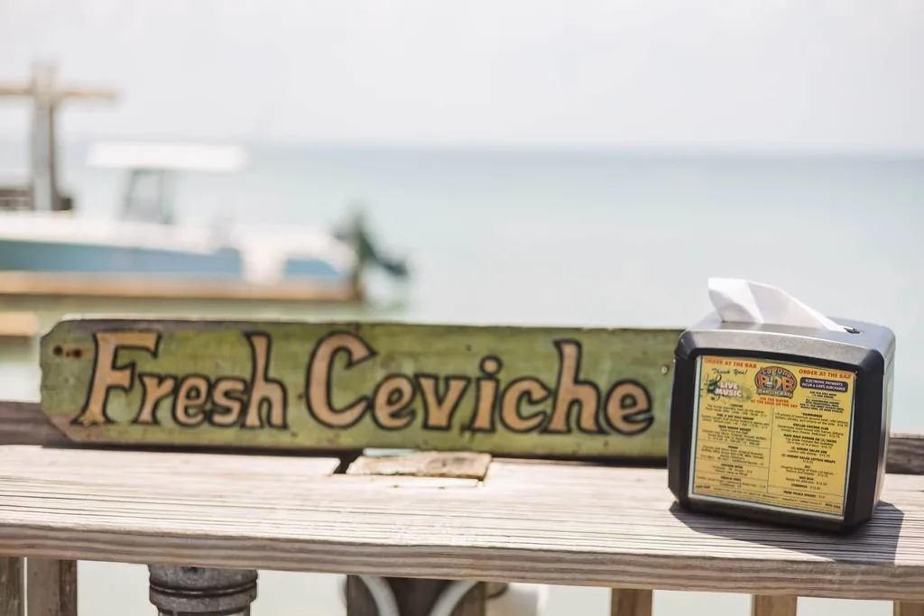 A hand-painted Fresh Ceviche sign on the bayside rail next to a napkin holder with the menu card, a boat behind