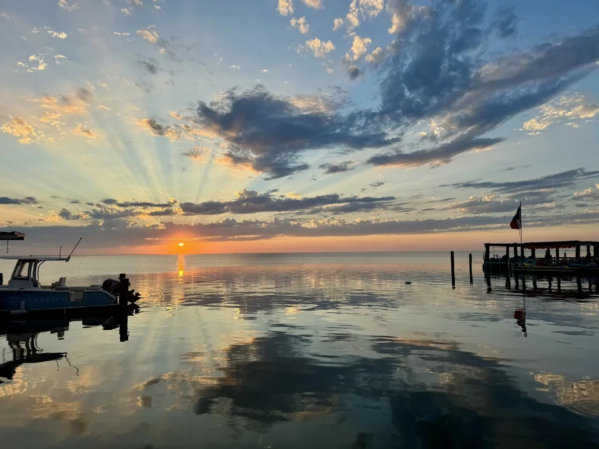 Sun rays through the clouds at sunset, reflected in calm water, with a boat and a pier