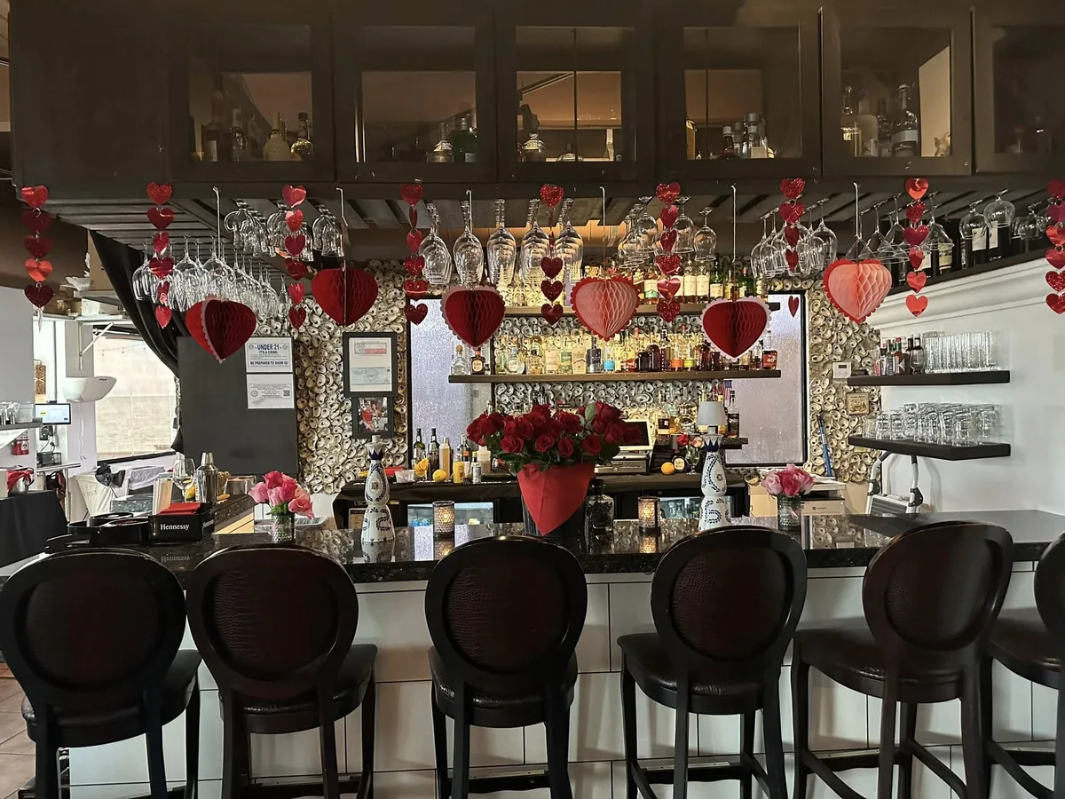 The bar with high-back stools, an oyster-shell wall behind the bottles and hanging wine glasses, decorated with red hearts
