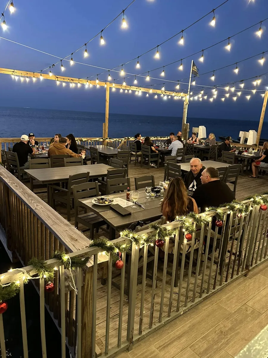 The waterfront deck at dusk under strings of lights, with diners at tables and the bay behind