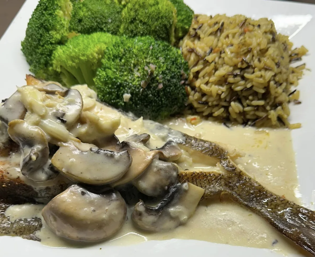 A skin-on fish fillet under mushrooms in a cream sauce, with broccoli and wild rice