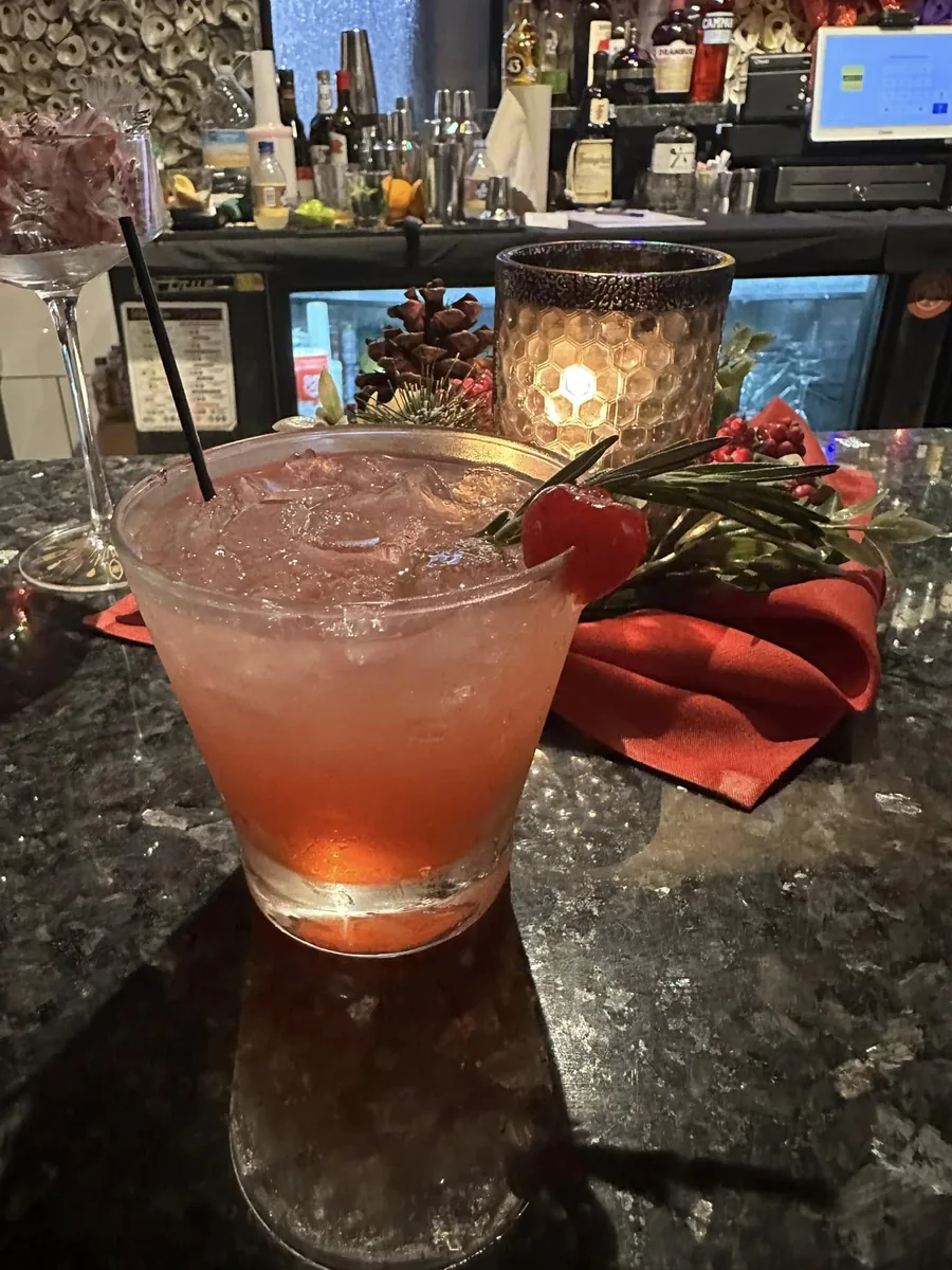 A pink cocktail on ice with a cherry and a rosemary sprig on the granite bar