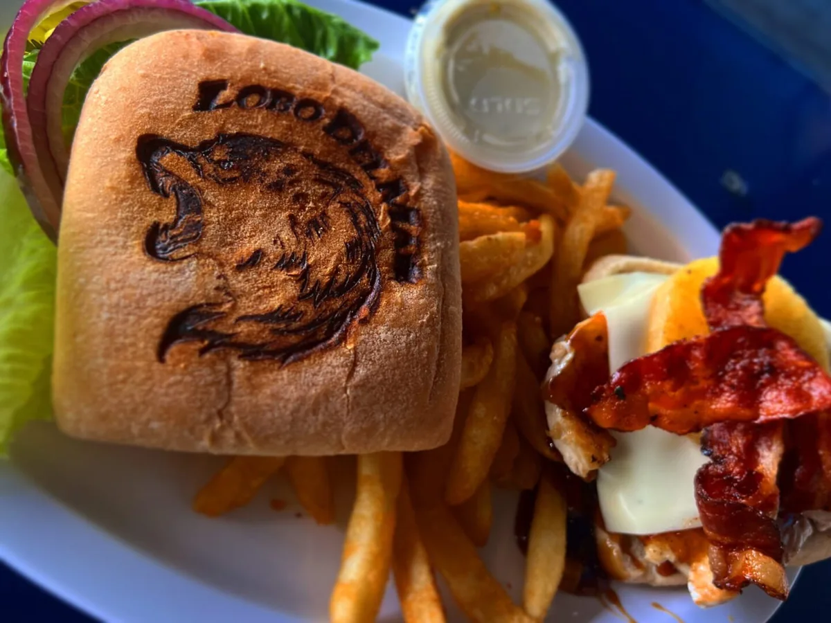 A sandwich on a bun stamped with the Lobo Del Mar wolf, with fries, bacon and melted cheese