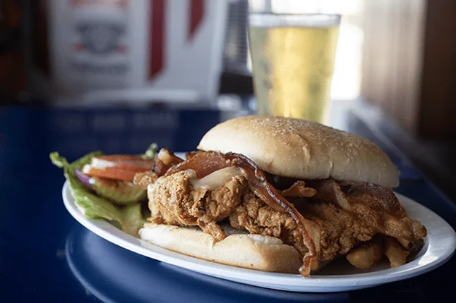 A fried chicken sandwich with bacon on a sesame bun, with lettuce and tomato and a glass of beer behind it