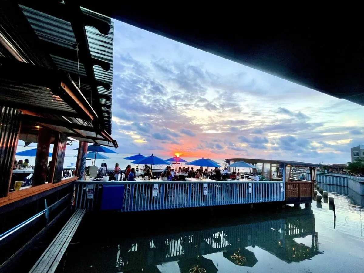 The sun setting over the bay beyond the deck and its blue umbrellas, reflected in the water