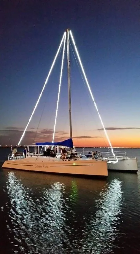 A catamaran with its rigging lit up, moored on the bay at dusk
