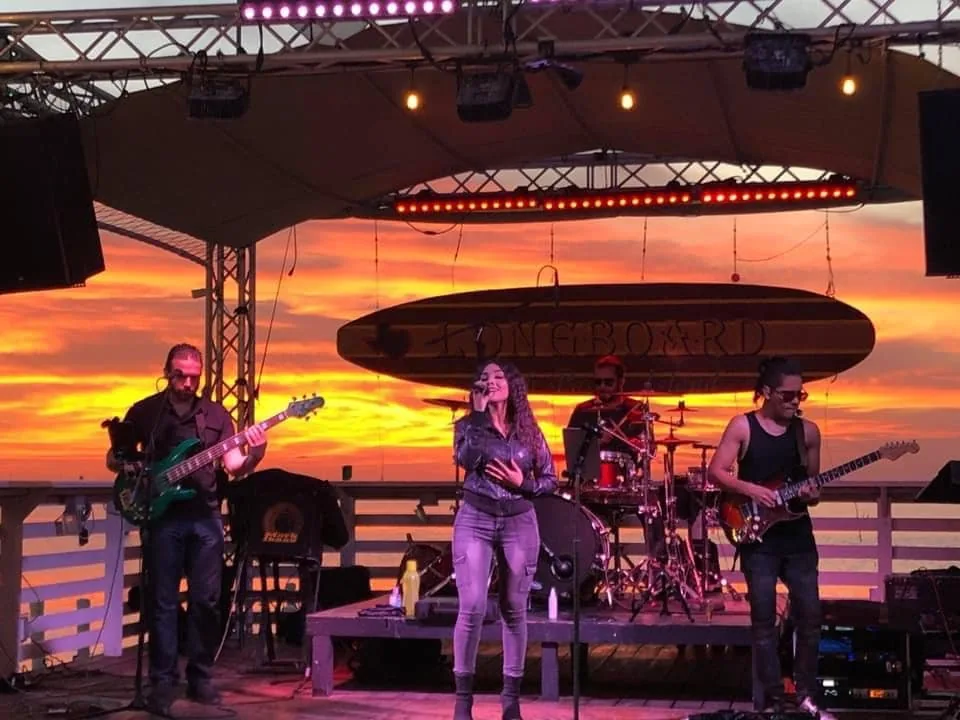 A live band playing on the stage under the Longboard surfboard sign at sunset