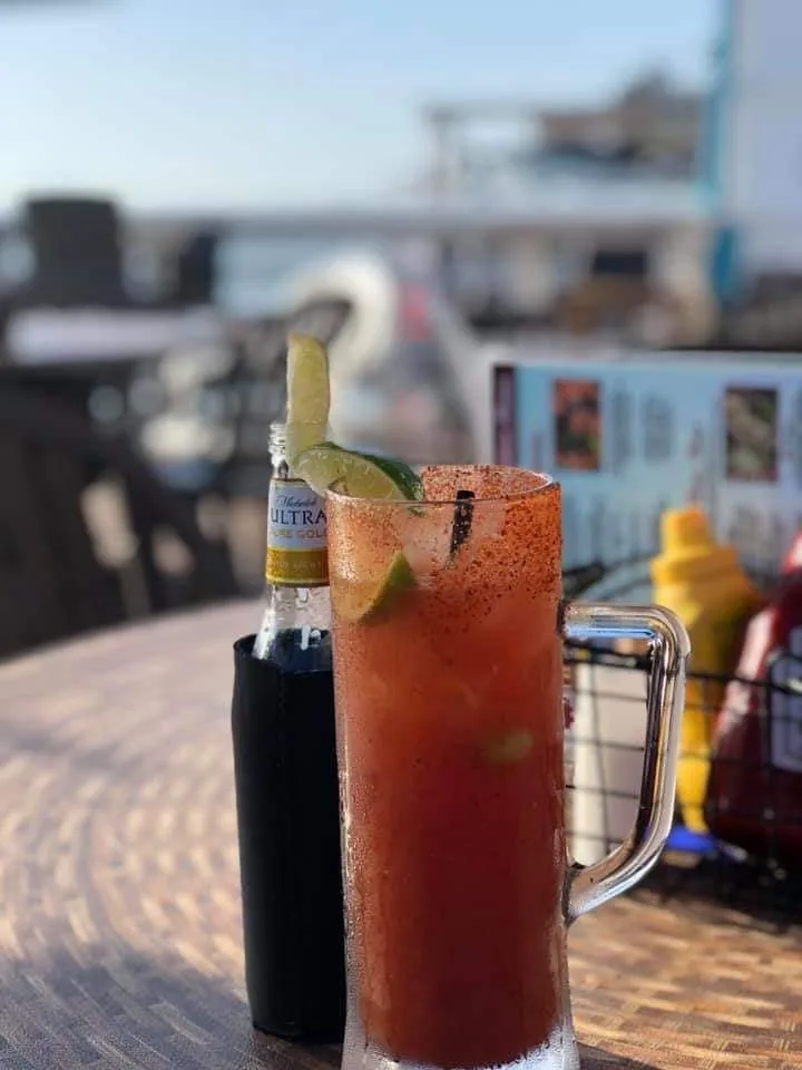 A michelada in a chili-rimmed mug with lime, next to a Michelob Ultra, on a table by the water