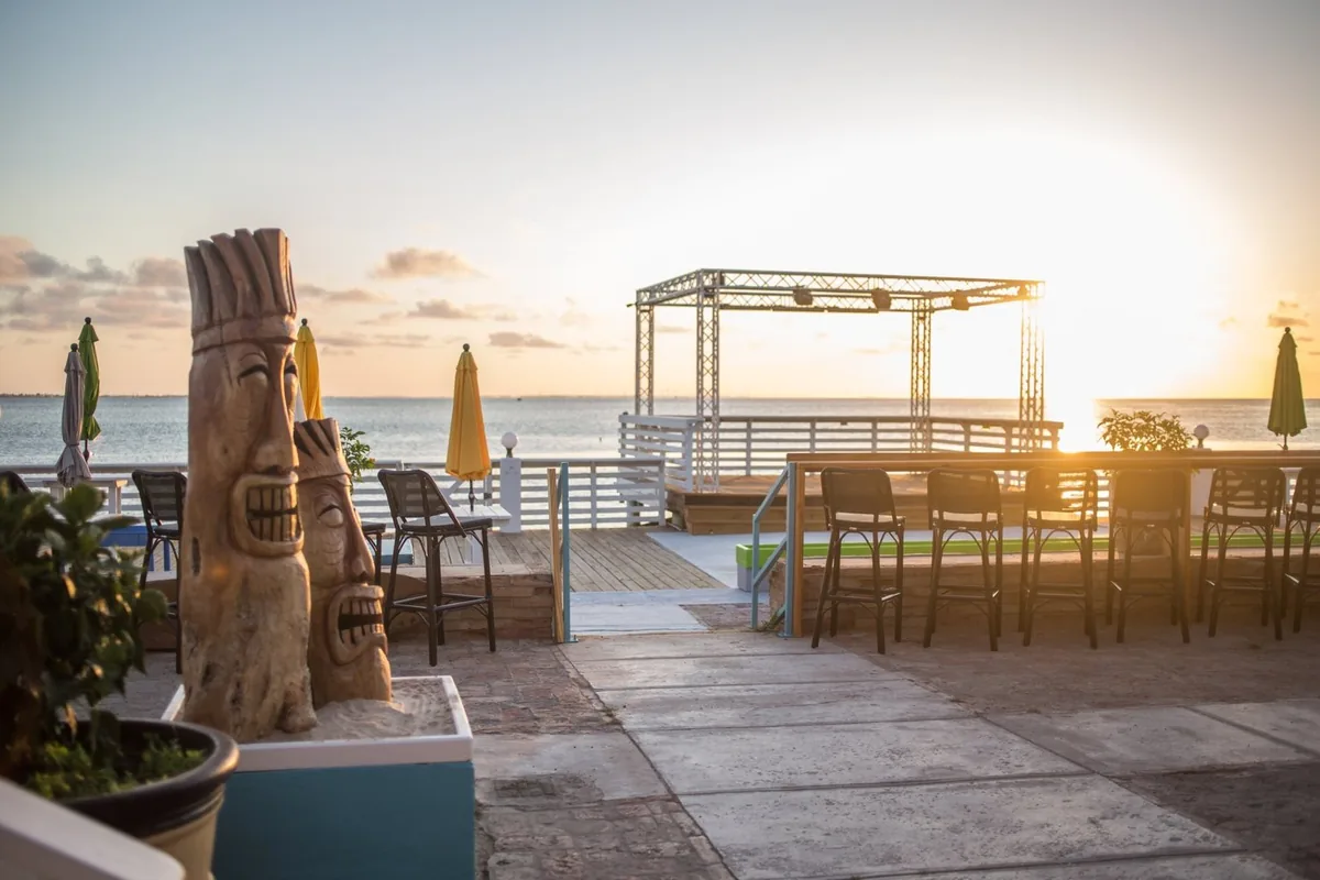 Carved tiki statues on the deck, with bar tables and the stage looking out over the bay at sunset