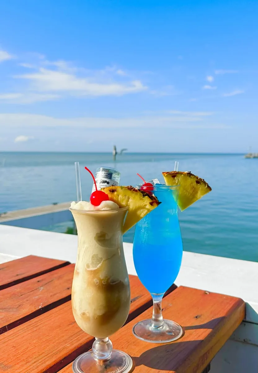 A frozen pina colada-style drink and a blue cocktail, both with pineapple and cherries, on the deck rail over the bay