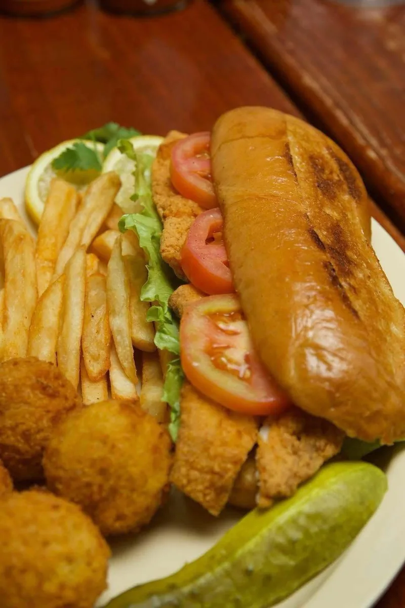 A fried seafood po-boy with lettuce and tomato, served with fries, hush puppies and a pickle spear