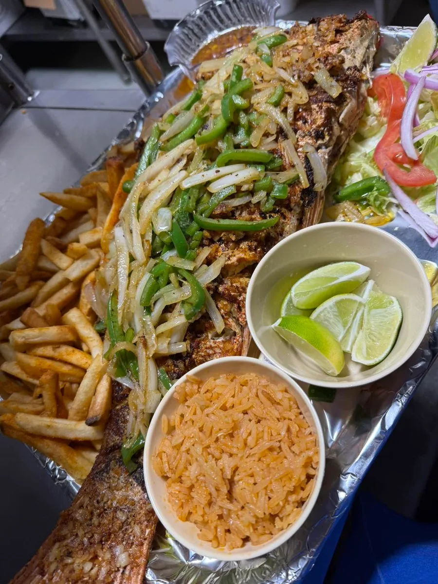 A whole fried fish topped with grilled onions and peppers, with fries, rice, lime and salad