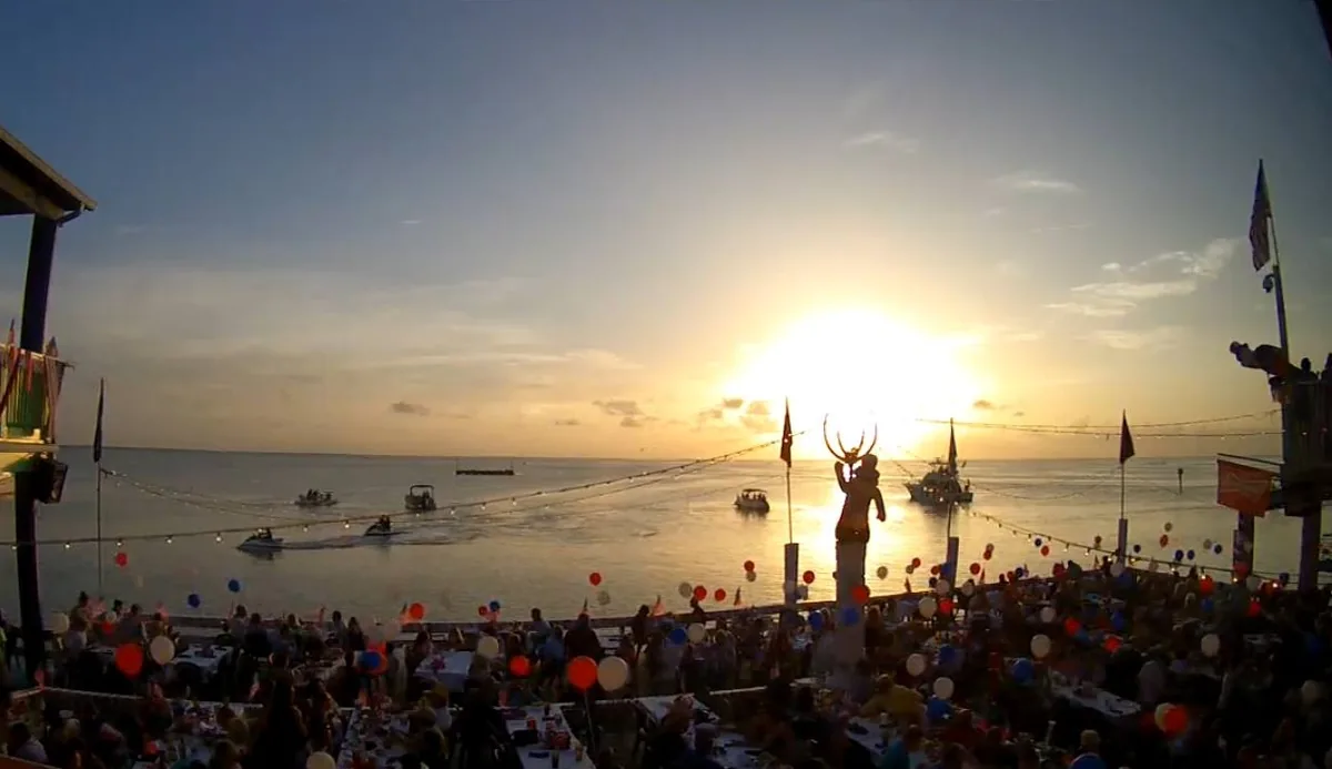 The sun setting over the bay, seen over a crowd on the deck with boats on the water