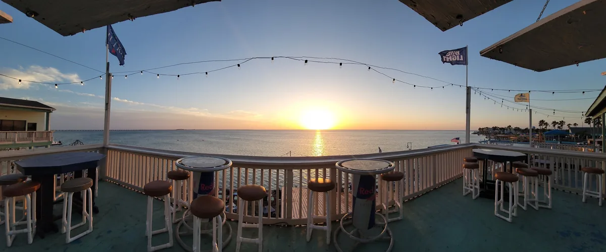 A wide view of the bay-front patio at sunset, with bar tables and stools along the rail and string lights overhead