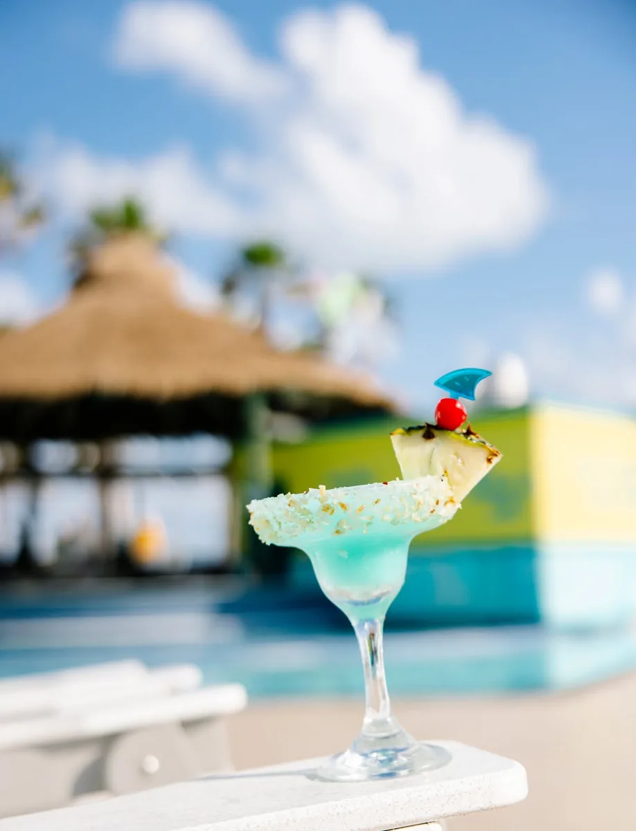 A blue margarita with a pineapple wedge and a cherry in front of the thatched pool bar