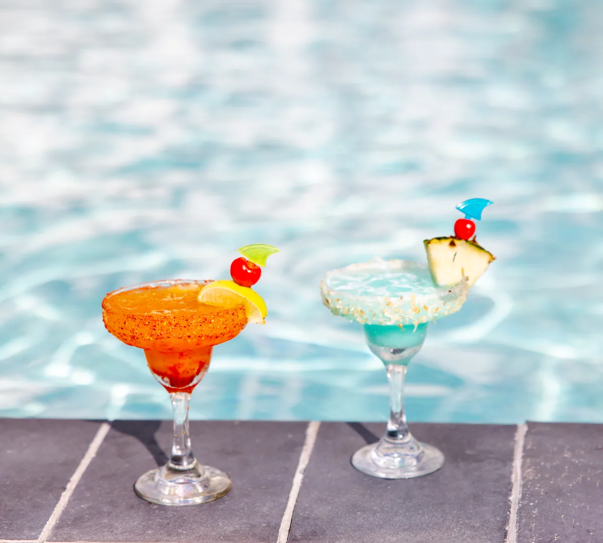 Two cocktails on the pool edge: an orange margarita with a chili-salt rim and a blue drink with a pineapple wedge and cherry