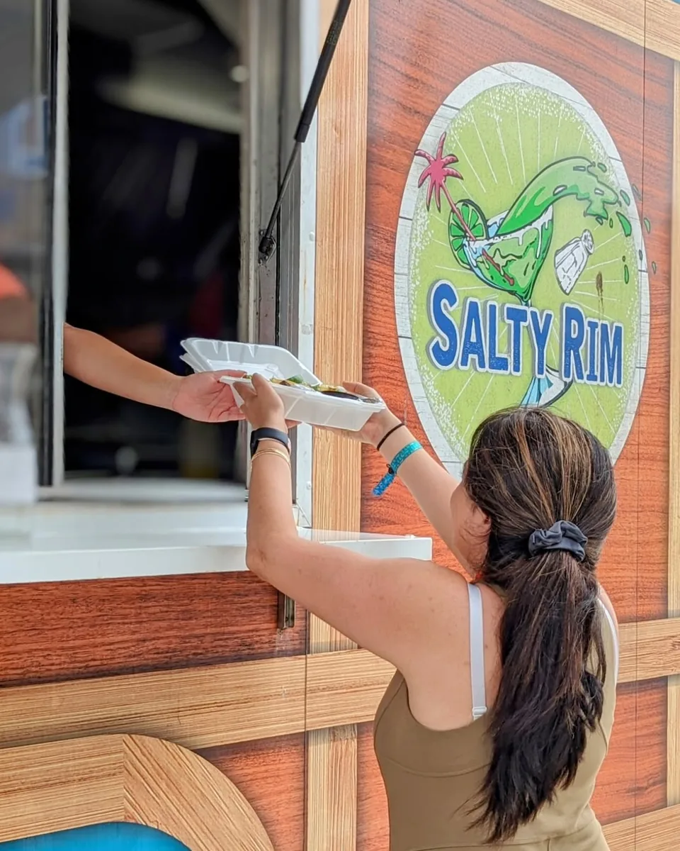 A guest, seen from behind, taking a takeout box at the Salty Rim's order window beside its painted sign