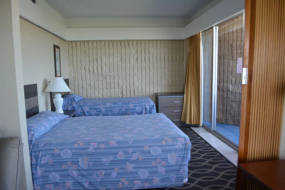 A room with two double beds in blue spreads and a sliding door to the balcony
