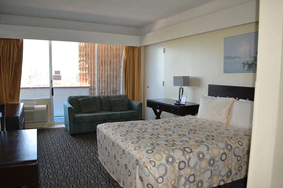 A room with a large bed, a sofa and a desk, and balcony doors letting in the light