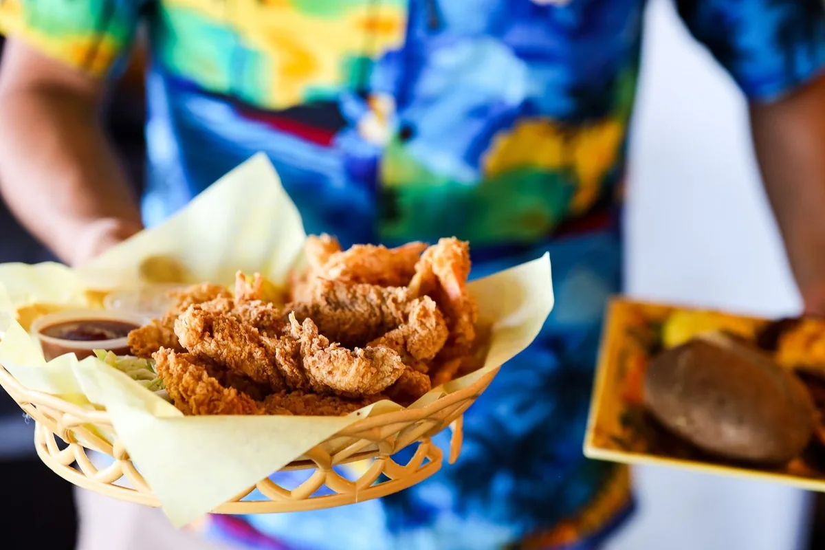 A server in a tropical shirt carrying a basket of fried shrimp and a plate with a baked potato