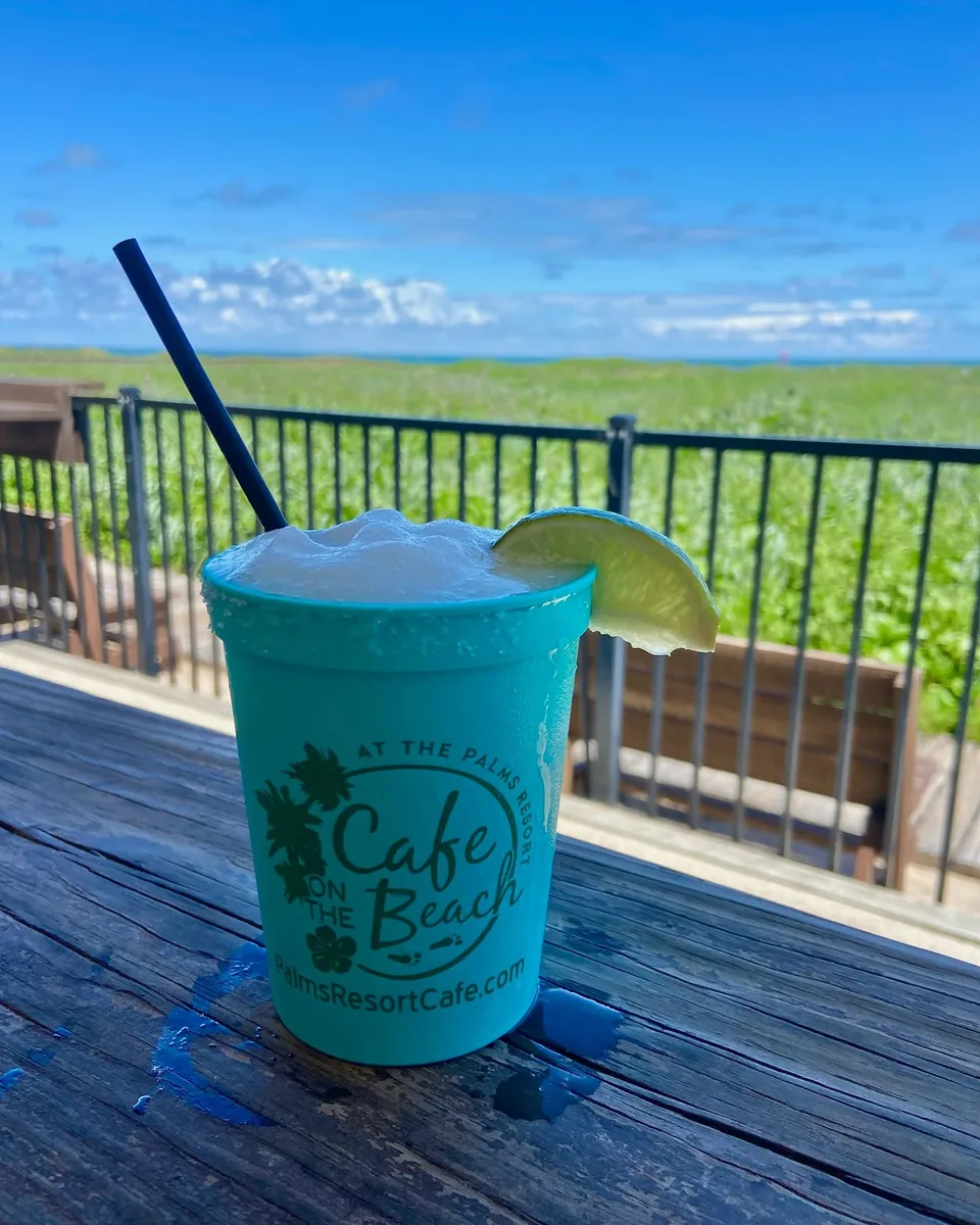 A frozen margarita with lime in a teal Cafe on the Beach cup on a table by the dunes