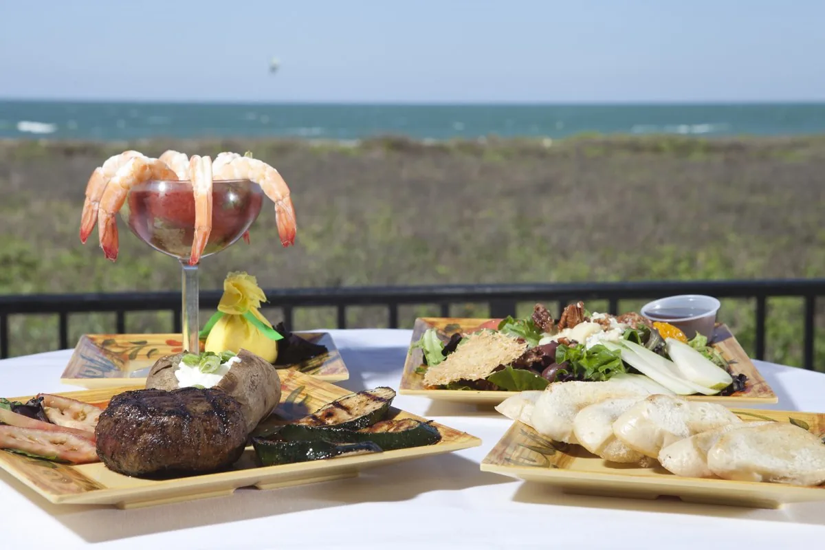 A table by the dunes with a shrimp cocktail, a steak with a baked potato and grilled vegetables, and a salad
